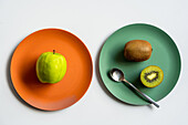 Von oben frischer leckerer grüner Apfel und reife ganze und halbierte Kiwi auf zwei farbigen Keramiktellern mit Silberlöffel auf weißem Tisch platziert