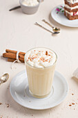 Glass of milk punch with cinnamon powder on whipped egg white against cake piece on cafeteria table on light background