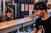 Asiatische Frau in Freizeitkleidung sitzt am Tresen und unterhält sich mit einem männlichen Angestellten einer modernen Ramen-Bar