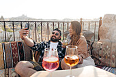 Positives, junges, stilvolles Paar, das auf einer Couch sitzt und mit einem Cocktail anstößt, während es ein Selbstporträt mit dem Handy auf einer Terrasse in Kappadokien, Türkei, macht