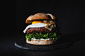 Leckerer Burger mit Ei auf dem Brötchen und frischem Salat auf einer Schiefertafel auf schwarzem Hintergrund im Studio serviert