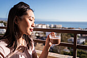 Glückliche Asiatin mit einem Glas Heißgetränk in der Hand auf dem Balkon sitzend