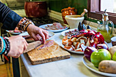 Abgeschnittener, nicht erkennbarer männlicher Koch in Uniform schneidet Fischfilet auf einem Brett, während er an einem Tisch mit verschiedenen Zutaten in der Küche steht