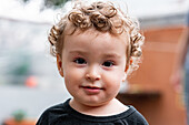 Entzückendes kleines Kind mit blondem, lockigem Haar und braunen Augen, das in die Kamera schaut, vor einem unscharfen Hintergrund