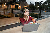 Durch das Glas einer jungen Bloggerin, die mit einer gesunden Mahlzeit am Tisch sitzt und einen Laptop benutzt, während sie Zeit in einem modernen Restaurant verbringt