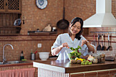 Happy asian woman in casual clothes stirring salad with assorted veggies and fruits with wooden spoon in bowl while cooking in kitchen on sunny day
