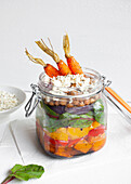 Salad with colorful ripe chopped bell peppers and bulgur topped with raw carrots served in jar on table against white background