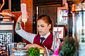 Seriöse Barkeeperin in Uniform steht am Tresen einer Bar und gießt Alkohol in einen Shaker, während sie einen Cocktail zubereitet