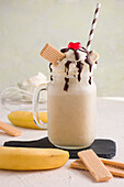 Glasgefäß mit süßem Bananensplit-Milchshake mit Schlagsahne, Waffeln, Schokolade und Kirsche auf Schneidebrett