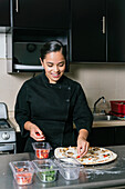 Positive ethnic female chef in uniform adding fresh cherry tomatoes on pizza while cooking at kitchen counter in modern restaurant