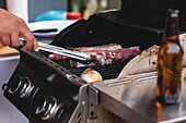 Abgeschnittene, nicht erkennbare männliche Person in Schürze, die rohes, mariniertes Fleisch auf den Grillrost eines Grillofens legt, während sie Steaks zubereitet