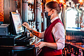 Seitenansicht einer Barkeeperin mit Schutzmaske, die eine Kasse bedient und den Bildschirm eines Displays berührt, während sie in einer Bar arbeitet