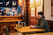 Zufriedene asiatische Frau in legerem Pullover, die an einem Holztisch in einer Ramen-Bar sitzt und mit einem breiten Lächeln in die Ferne schaut