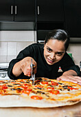 Focused young ethnic female chef in uniform cutting fresh cooked pizza with blade roller