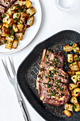 From above of appetizing fried beef chop with potatoes topped with minced garlic and parsley served on frying pan and plate on white background with cutlery