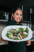 Young ethnic female chef in uniform holding plate with fresh delicious salad of corn vegetables pears