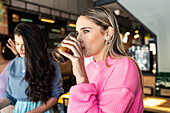Side view of trendy young female in fashionable outfit drinking glass of cold coke and looking away while spending time with friends in cafe
