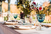 Nahaufnahme einer gedeckten Festtafel mit Kristallgläsern, Besteck und Serviette auf einem Teller neben einem Strauß frischer Blumen für eine Hochzeit und einer Menükarte
