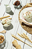 Leckeres Eis in Schalen mit Waffelröllchen und Waffeltüten auf weißem Holztisch in der Nähe von Krügen mit Milchschokoladenriegeln und Walnüssen von oben
