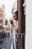 Side view of reflective African American female with cup of hot beverage looking at camera from balcony in city