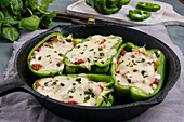 Halves of green bell pepper with pizza stuffing and melted cheese in skillet pan on table with green herbs in kitchen