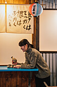 Seitenansicht einer überglücklichen Asiatin im Pullover, die an einem blauen Tisch mit einem traditionellen Getränk in einem Café sitzt
