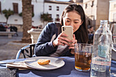 Smiling Asian female taking picture of tasty fired snack on cellphone near female friend while sitting at table in outdoor cafeteria