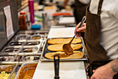 High angle side view of crop anonymous male cook with ladle spreading sauce on baked flatbread in kitchen of restaurant