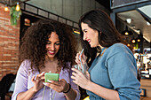 Lächelnde junge Freundinnen in Freizeitkleidung, die bei einer Limonade im Restaurant auf ihrem Handy surfen