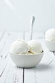 Coops of vanilla ice cream served in white ceramic bowls with spoons near waffle cones scattered on white table