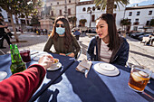 Asian woman near female in medical mask sitting at table with crop anonymous friend in outdoor cafeteria during coronavirus pandemic