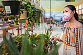 Young ethnic female buyer in disposable mask choosing potted plants while looking away in garden shop