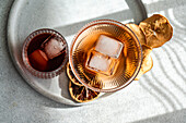 Draufsicht auf zwei Gläser, eines mit Kirschlikör und Eis, das andere mit einem rosigen Likör, daneben goldene getrocknete Zitrusfrüchte, auf einem runden Steintablett