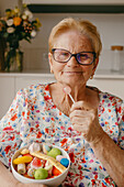 Eine fröhliche ältere Dame mit Brille lächelt, während sie einen rosa Lutscher und eine Schale mit bunten Bonbons in der Hand hält