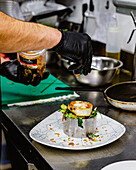 Ein Koch in einer Großküche gibt einem Gourmetteller mit grünen Kräutern und Toppings den letzten Schliff