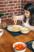 A focused young girl adds cheese to her tortilla, preparing a taco at a wooden table with ingredients laid out for a family meal