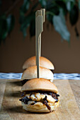 Three mini beef burgers topped with melted cheddar cheese served on a rustic wooden plank, skewered for presentation.