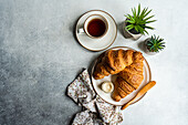 Draufsicht auf eine Frühstücksszene mit flockigen Croissants, einem Glas Honig, einer Tasse Tee und dekorativen Sukkulenten in Töpfen auf einer strukturierten Oberfläche