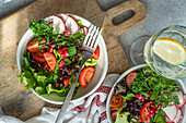 Top view of close up of a healthy vegetable salad with mixed greens, radishes, cherry tomatoes, and pomegranate seeds, next to a glass of lemon water on a wooden board