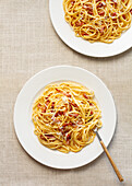 Draufsicht auf klassische Spaghetti Carbonara, garniert mit Käse und Speckstückchen, serviert auf weißen Tellern mit rustikalem Hintergrund.