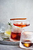 A trio of elegantly served alcoholic drinks in textured glasses, garnished with a cinnamon stick and fresh rosemary, displayed on a stone platter with a soft backdrop