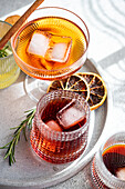 An array of exquisite alcoholic beverages in decorative glasses, accompanied by ice cubes, a cinnamon stick, rosemary, and dried citrus slices on a marbled tray