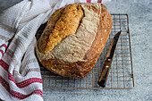 A freshly baked loaf of rye sourdough bread sits cooling on a wire rack, accompanied by a bread knife and a white cloth with red stripes