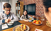 A family enjoys a weekend breakfast together at home with a spread of fruits, muffins, cookies, and cereal on the table