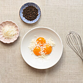 Draufsicht auf rohe Zutaten für Spaghetti Carbonara, darunter Eier, geriebener Käse, Pfefferkörner und ein Schneebesen auf einer Stoffoberfläche.