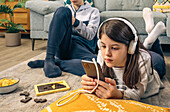 A focused girl with headphones uses a smartphone while lying on a floor cushion, with her brother in the background and a puzzle on the side