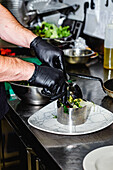 Nahaufnahme der Hände eines Kochs mit schwarzen Handschuhen, der einen Gourmetsalat in einem Metallring auf einer marmorierten Platte in einer Profiküche anrichtet