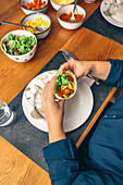 Anonymous hands carefully hold a filled taco over a plate, with bowls of toppings like lettuce, cheese, and sauce on the dining table