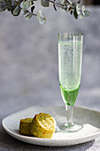 A glass of sparkling pistachio vodka next to a pistachio cookie, elegantly displayed on a white plate with eucalyptus in the background, set on a concrete surface