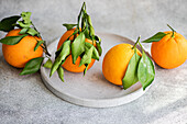 A trio of ripe oranges adorned with lush green leaves, presented on a grey circular stone plate against a textured background